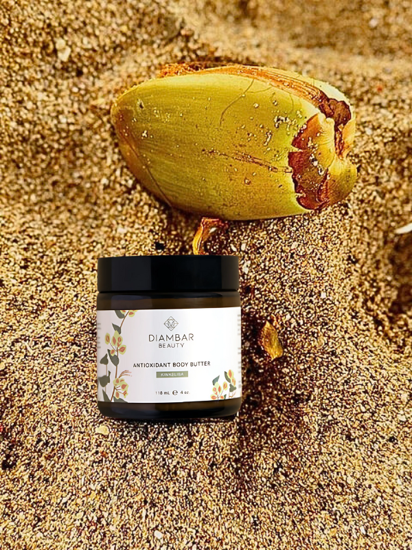 A beautiful jar of diambar beauty kinkeliba body butter laying in a bed of sand alongside a fresh coconut from Senegal
