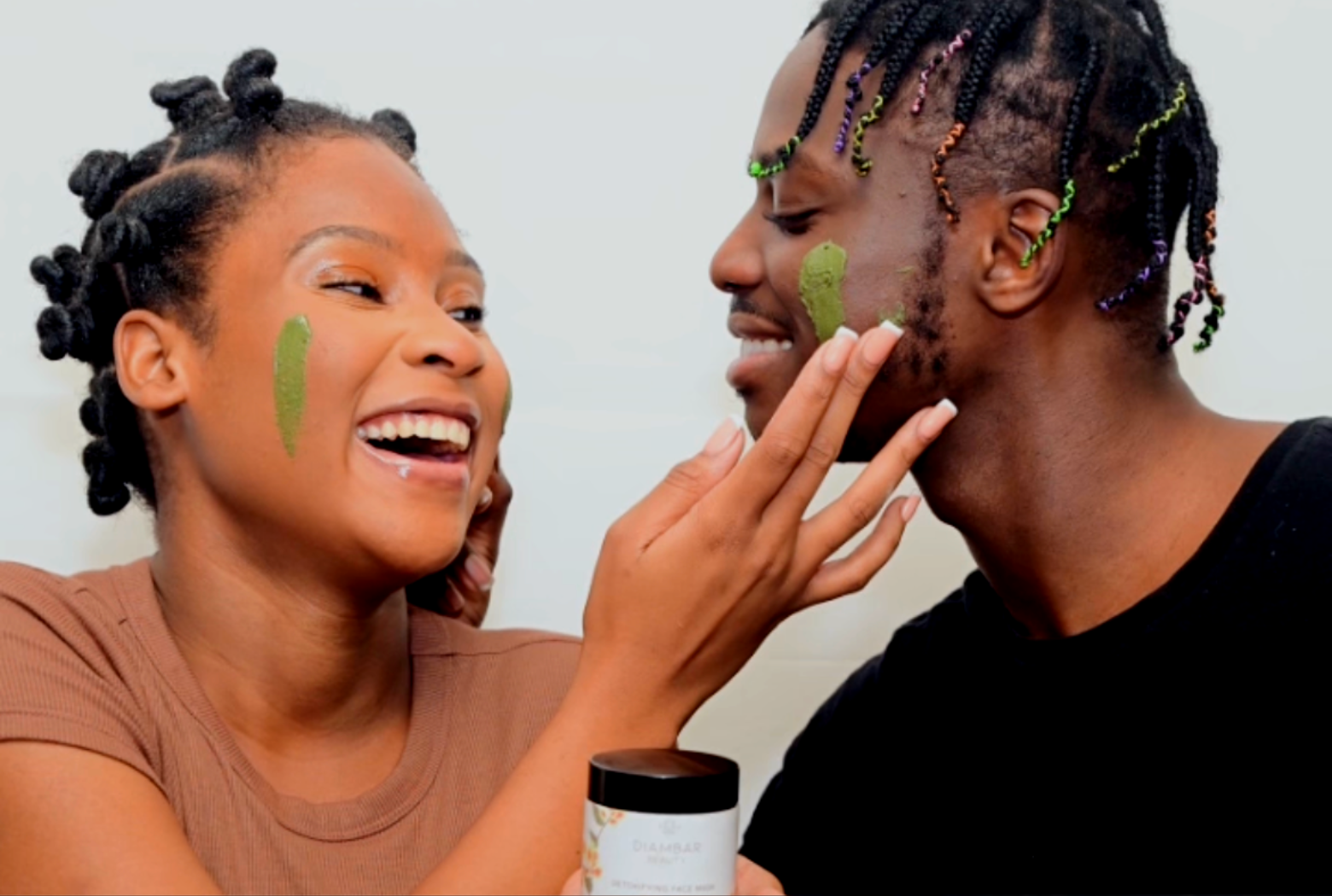 A joyful man and woman smiling at each other while playfully swiping the green Kinkeliba Detox Face Mask onto each other's cheeks.