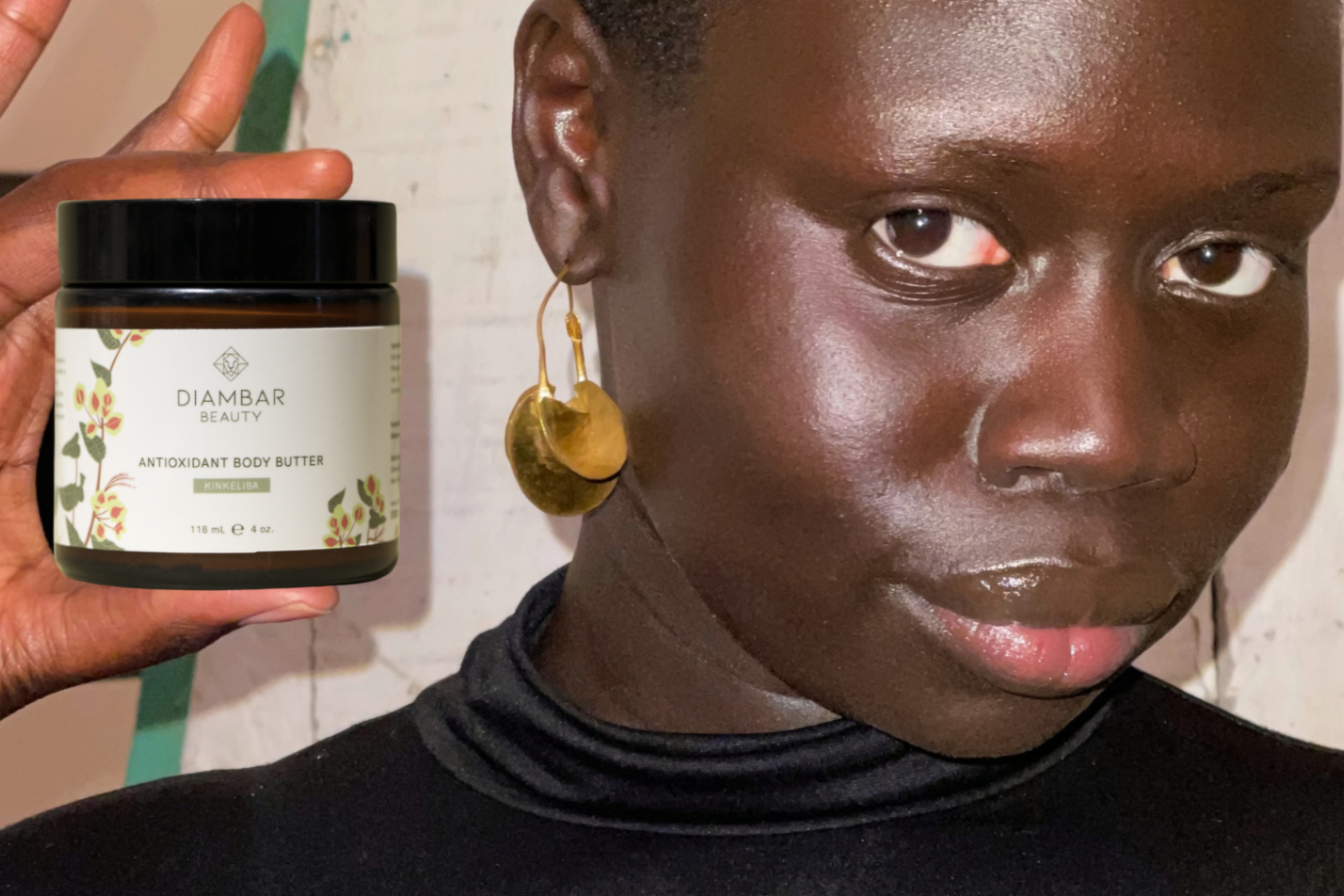A radiant dark-skinned woman holding up a glass jar of Kinkeliba Antioxidant Body Butter, showcasing its luxurious packaging.