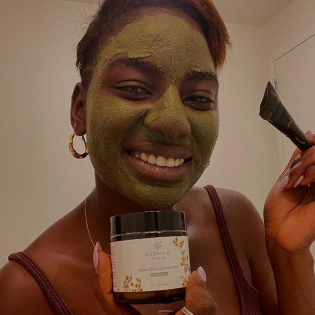 woman with the green Kinkeliba Detox Face Mask applied, smiling as she holds up the mask jar in one hand and a mask brush in the other.